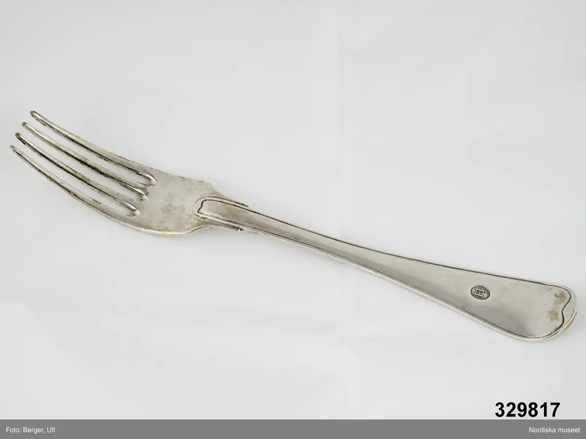 Fyrkloig matgaffel,silver, enkelräffla, svagt uppsvängt rund ända. Monogram på framsidan. Klack vid roten. Stämpel på baksidan: N2 - kattfot - St Erik- Zethelius. Baksidans räffla spadlik vid skaftroten.

Ovalt monogram, pärlstavskant. Versalt H antikva med förnamnsbokstaven J sammanfallande med första H-stapeln,  J-prick ovanför stapeln.

Har tillhört Lärftskramhandlare Johannes Hazelius, Stockholm. Se även inv.nr. 329.815 (stämpel)  och 329.816 (gaffel märkta med denna stämpel).
/2007-03-19 Heidi Henriksson