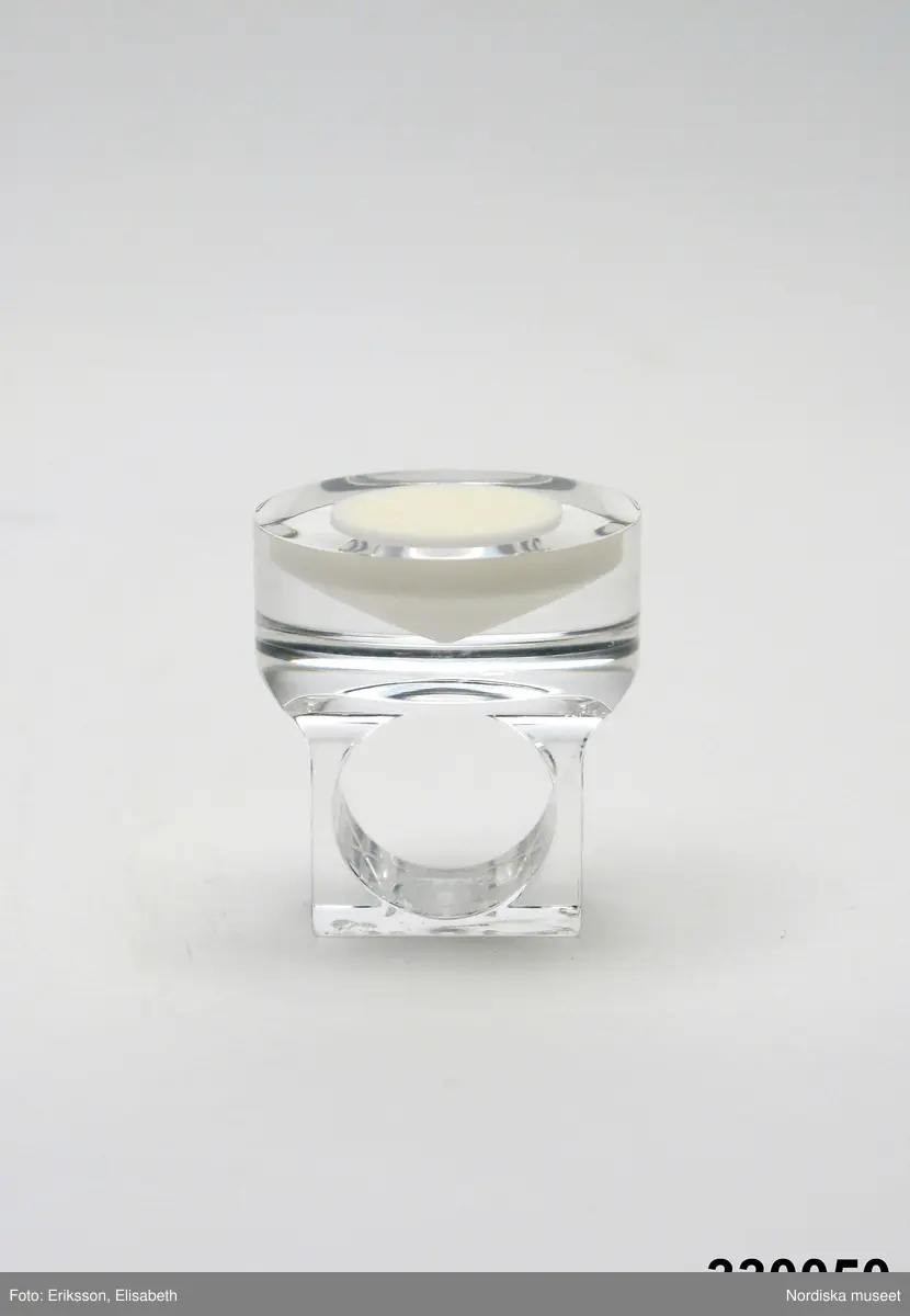 Ring av transparent akrylplast med konformig inläggning av vit plast. Cirkelrunt framstycke, skena med yttre kvadratisk form. Designad av silversmeden Siv Lagerström under 1960-talets senare del. Tillverkad av Gravyrverken i Sollentuna (råämne som kapades och frästes ur) och Siv Lagerström (urborrning för fyllning av färgad Castolite, en form av gjutplast). Inskription på skenans bakre del "Siv" samt "MADE IN SWEDEN". 
/Ingrid Roos 2010-04-07