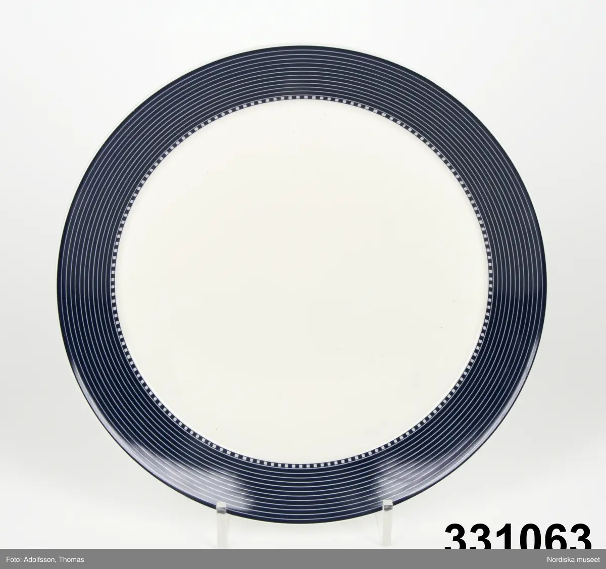 Mattallrik, modell Teema, stengods. Tillverkad av Arabia, design Kaj Frank. Vit och blå tallrik, med mörkblå hög utåtlutande kep med smala vita linjer samt vid mötet mellan kep och spegel dekor av vita och blå kvadrater. Vit  odekorerad spegel. Inget bräm.
Stämplad under tallriken.

/Cecilia Wallquist 2011-07-19