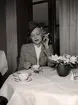 Modebilder, från Nordiska Kompaniet 1940. En kvinna klädd i dräkt och hatt sitter vid ett bord. Framför henne står en kopp kaffe, hon håller en cigarett i handen.