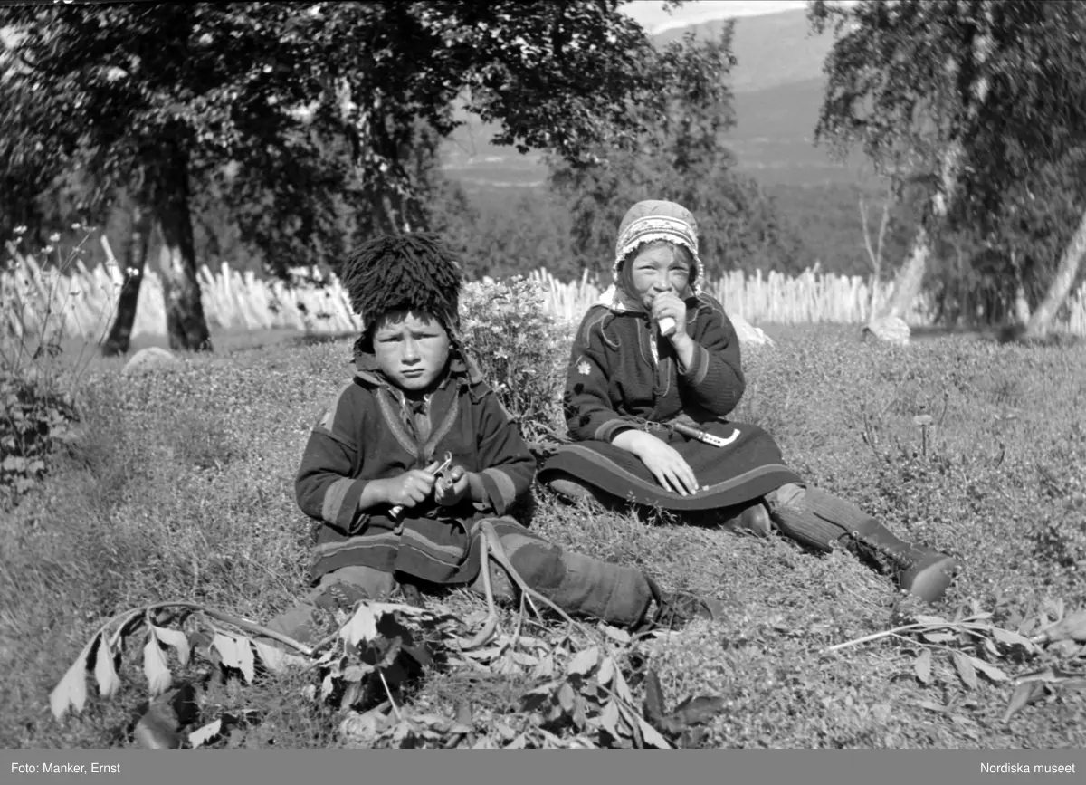 Två barn sitter utomhus och äter kvanne. Vilasund, Umbyns sameby. Artfjället.

Text på ark i arkivet: "Soutsgärdan / Kauresando-lapska barn, gosse och flicka, skala och äta kvannestjälkar; barn till Olof Omma."