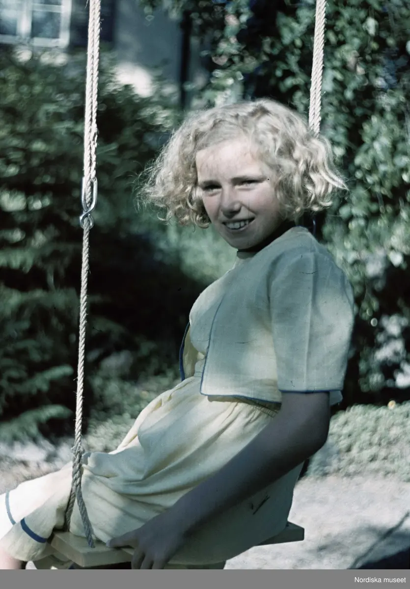 Flicka i gul klänning med bolero vid gunga. Troligen fotograf Gunnar Lundhs dotter Jytte.