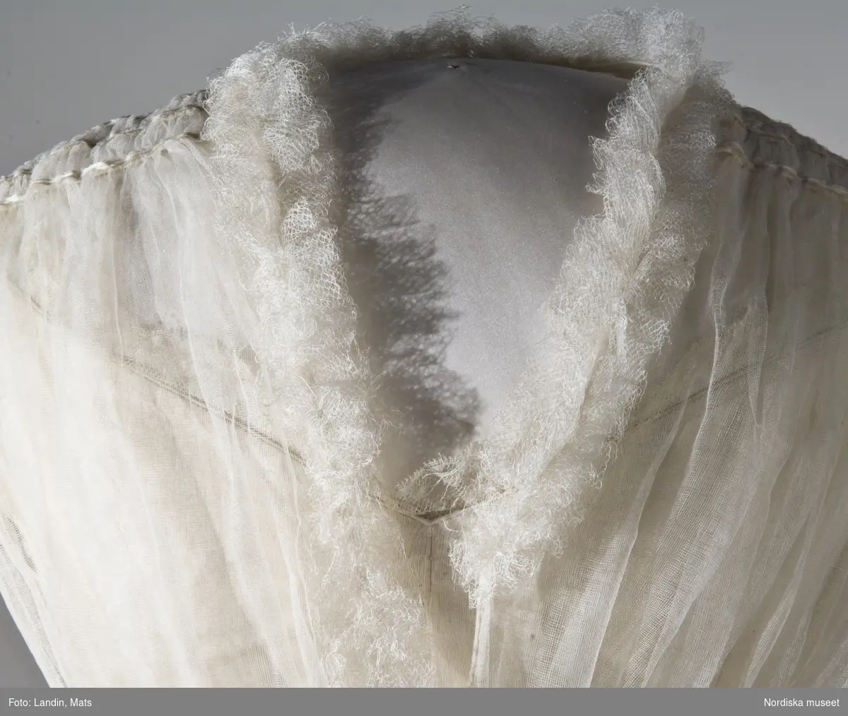 Brudklänning, liv med fastsydd kjol, vit tarlatan, garnerad med vita sidenband, rosetter. Klänningen har ett löst skärp av vit tarlatan.