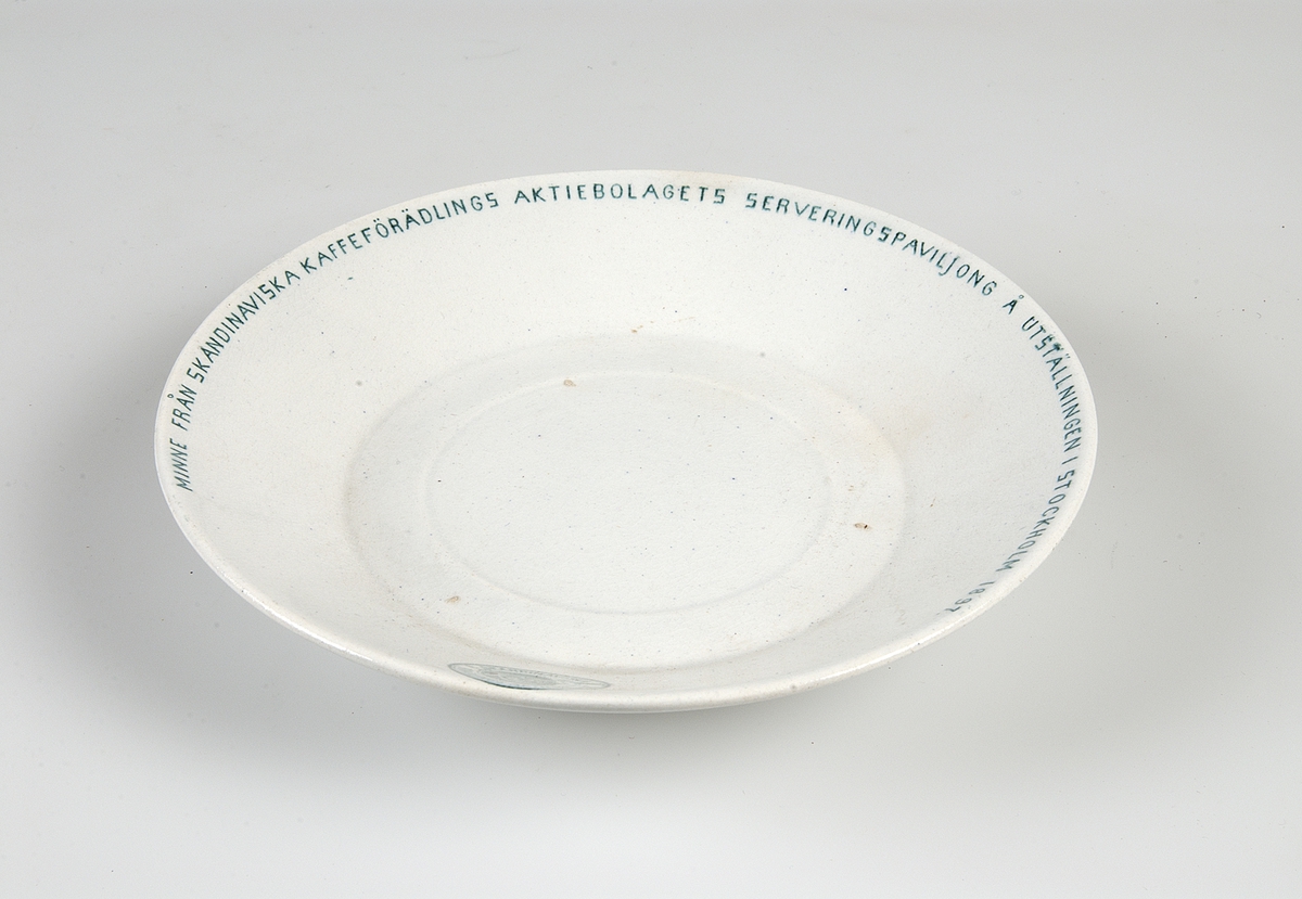 Kaffekopp med fat, vitglaserade med grön dekor. Runt kanten: "Minne från skandinaviska kaffeförädlings aktiebolags serveringspaviljong å utställningen i Stockholm 1897". Samma text på fatet. Grön tryckt stämpel: "GUSTAFSBERG".