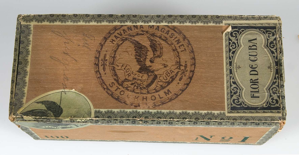 Papperslister i grönt och svart. Text på lockets pappersetiketter: Flor de Cuba. På locket en stämpel med texten: Havanna Magasinet Stockholm; Flor de Cuba, samt en örn. Blyertsanteckningar på locket: Till julgran. På framsidan i grönt tryck: 100, No 1. Fodrad med grönt papper. På lockets insida ett byggnadsmotiv i svart, på skyddsbladet en örn i svart.