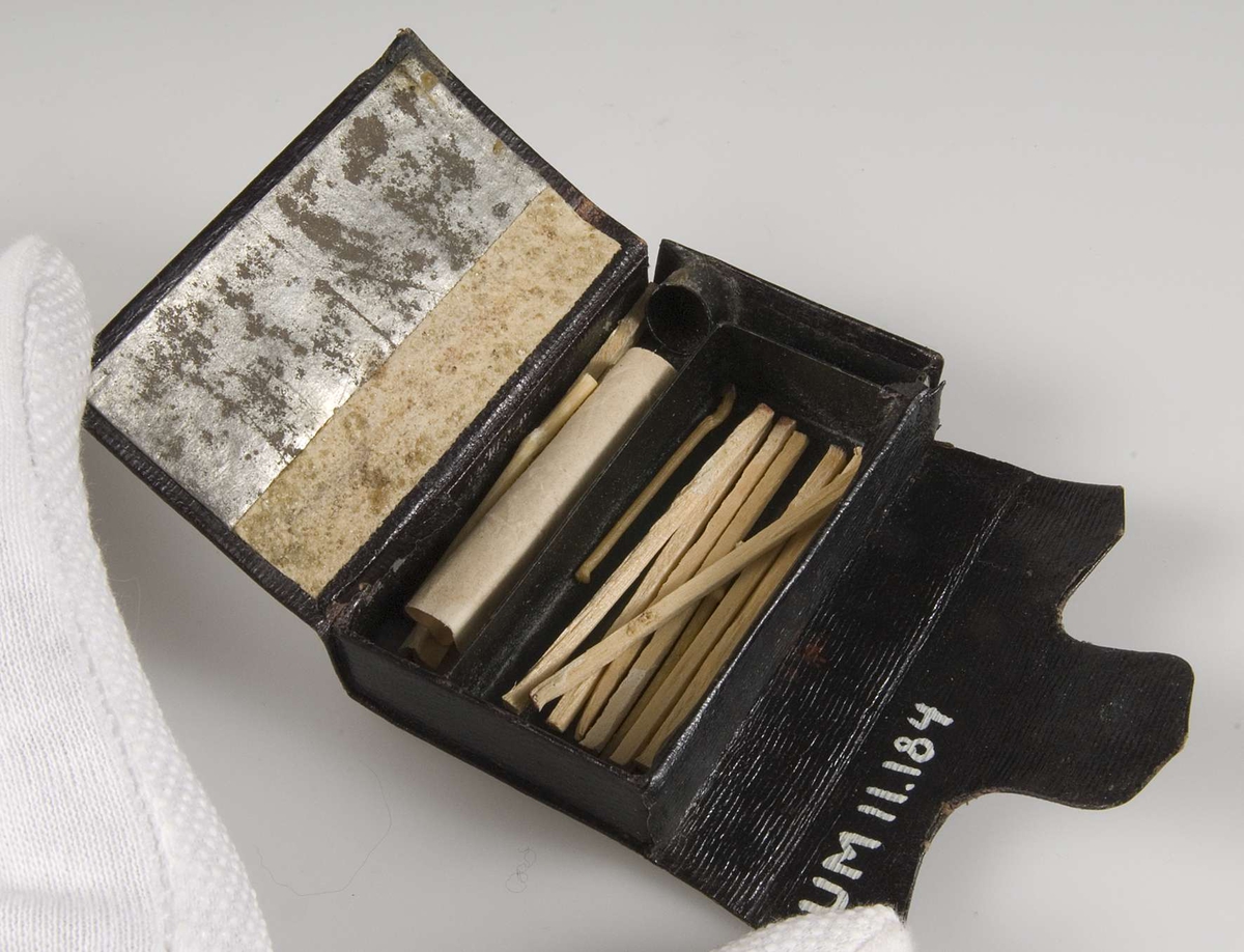 Ask av brunt läder med framsida av pärlbroderi på perforerad papp. Pärlor i grönt, blått, vitt, silver och guld. Askstomme av plåt.