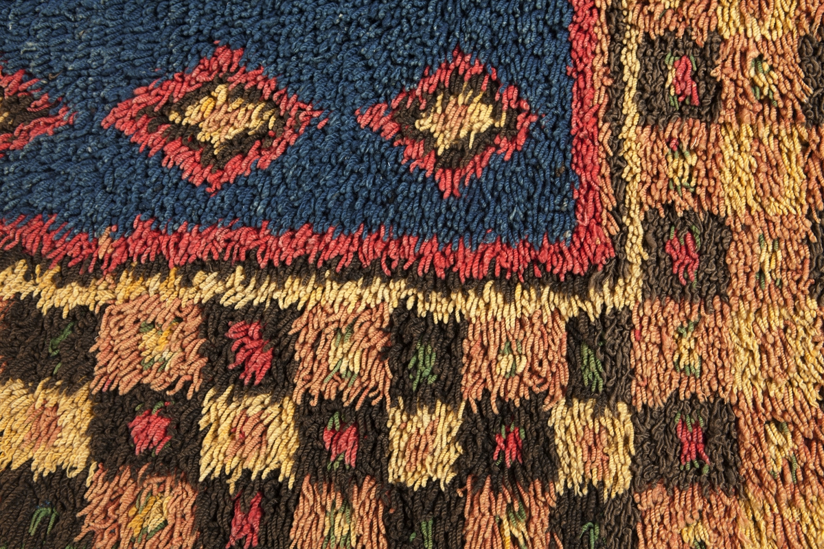 Prydnadsrya i två våder, hopsydda kant i kant. Botten i korndräll med varp av blått s-tvinnat lingarn 11 tr/cm och inslag av mörkbrun s-spunnen ull 7½ inslag/cm. Nockor av en tråd tvåtrådigt z-tvinnat hårdtvinnat ullgarn, knutna med giordesknut över 4 varptrådar med 4 varptrådar mellan nockorna och 4 inslag mellan nockraderna. Nockorna syns ej på baksidan.

Mönster: Stort blått mittfält avgränsat mot bården av en röd, en grönbrun och en gul smal linje. På det blå mittfältet, utmed sidorna romber av rött, grönbrunt och gult. Bred bård av schackrutor i grönbrunt, gult och rosagult. Gröna, röda och gula prickar i rutorna. Ytterst smal grön rand.