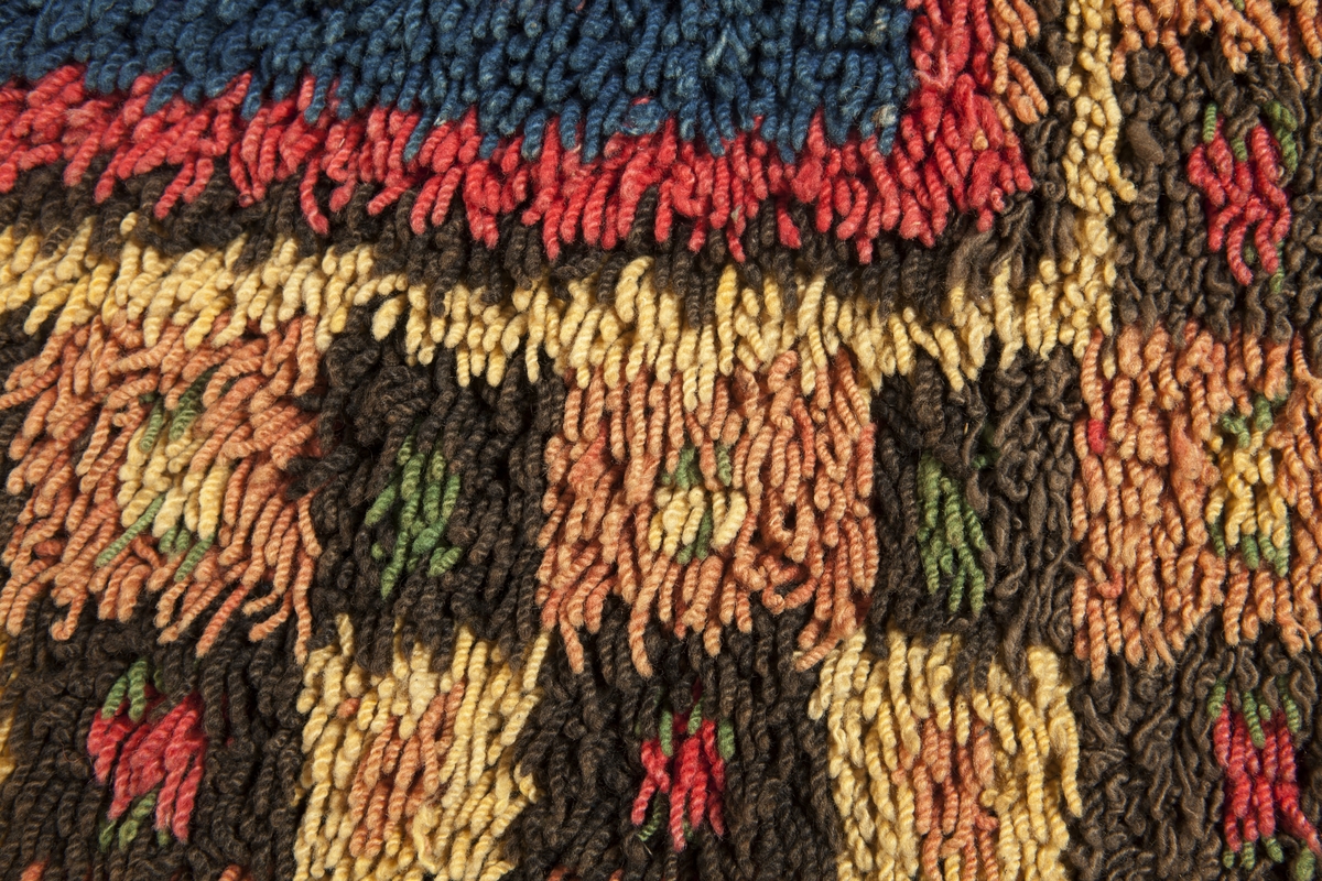 Prydnadsrya i två våder, hopsydda kant i kant. Botten i korndräll med varp av blått s-tvinnat lingarn 11 tr/cm och inslag av mörkbrun s-spunnen ull 7½ inslag/cm. Nockor av en tråd tvåtrådigt z-tvinnat hårdtvinnat ullgarn, knutna med giordesknut över 4 varptrådar med 4 varptrådar mellan nockorna och 4 inslag mellan nockraderna. Nockorna syns ej på baksidan.

Mönster: Stort blått mittfält avgränsat mot bården av en röd, en grönbrun och en gul smal linje. På det blå mittfältet, utmed sidorna romber av rött, grönbrunt och gult. Bred bård av schackrutor i grönbrunt, gult och rosagult. Gröna, röda och gula prickar i rutorna. Ytterst smal grön rand.