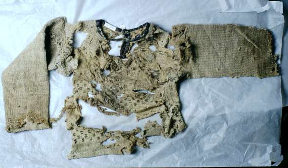 Fragment av stickad tröja med mönsterstickad bård i brunt och beige ullgarn. Bålen i 1-trådigt ullgarn mönsterstickad i rutmönster i brunt, natursvart och beige. Ärmarna är rundstickade i 2-trådigt ullgarn i naturvit, natursvart, brunt och beige. Sprund mitt fram 10 cm långt. Halsurringning och sprund mitt fram kantad med samma tyg som finns som lagningar på UM23252 och A 38 (UM ?). Hyska och hake i mässing mitt fram.
