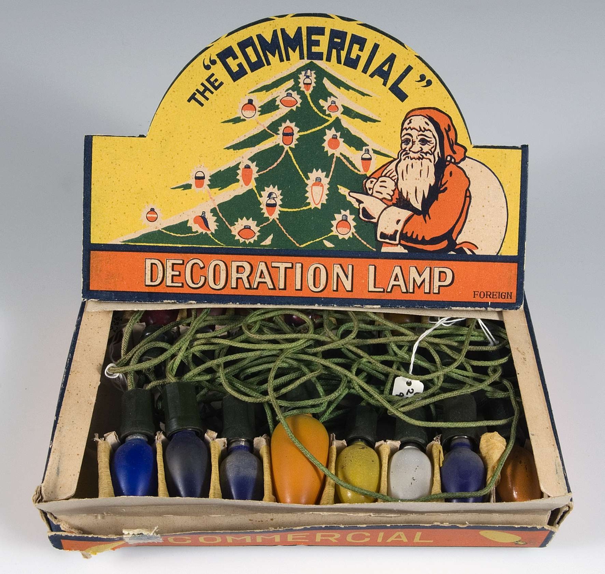 Pappask innehållande kulört elektrisk julgransbelynning, 16 små äggformade lampor av glas i blått, gult, vitt , rött, grönt och orange , i hållare av grön plast och med sladdar klädda med grön tråd. Några lampor är större än de andra och troligen utbytta. På askens lock bild av tomte och julgran i rött, gult, grönt och vitt och text: The Commersial Decoration lamp. Foreign.