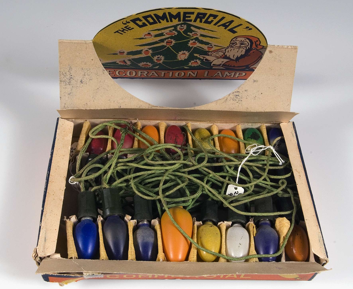 Pappask innehållande kulört elektrisk julgransbelynning, 16 små äggformade lampor av glas i blått, gult, vitt , rött, grönt och orange , i hållare av grön plast och med sladdar klädda med grön tråd. Några lampor är större än de andra och troligen utbytta. På askens lock bild av tomte och julgran i rött, gult, grönt och vitt och text: The Commersial Decoration lamp. Foreign.