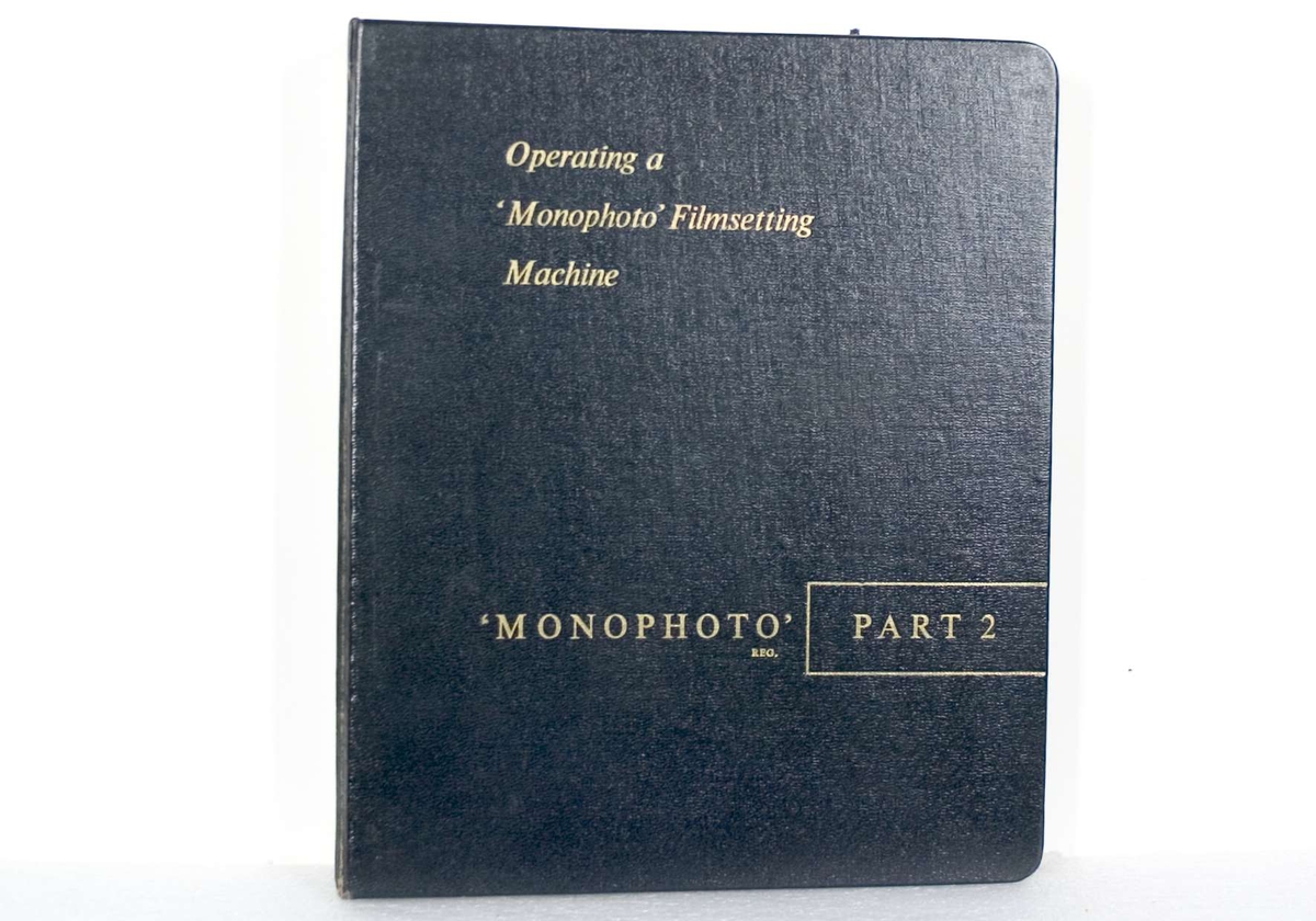 Handbok: Svart ringpärm av tjock papp och med metallinsats. Texten på utsidan är i guldfärg. Handboken har titeln "Operating a 'Monophoto' Filmsetting Machine, PART 2". På försättsbladet står bland annat "THE MONOTYPE CORPORATION LIMITED Registered Office: Monotype House, 43 Fetter Lane, London, E.C.4 Head Office and Works: Salfords, Redhill, Surrey". I pärmen ligger flera lösa blad, däribland några ritningar. Några lösa blad verkar ha varit fasttejpade på olika sidor i pärmen.