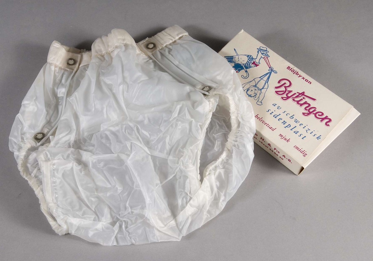 Blöjbyxa av plast i kartong av papper. Tryck i blått och rött, text om blöjbyxan, bland annat:  Blöjbyxan Bytingen av schweizisk sidenplast... samt David Grute H. & F:s A-B, Stockholm 15...