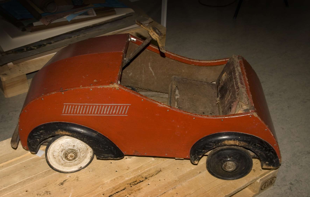 Trampbil, kaross av rödmålad masonit. Skärmar av svartmålat trä. Stötfångare fram av trä, med rester av ljust grå färg. Mönster i grått på båda långsidorna. Baktill en målad bilskylt med texten: AF-41. Bak även bokstaven S i svart. Tre svarta hjul av järn med däck av vitt gummi. Ett vitmålat hjul med däck av svart gummi. Sits av rött läder på stomme av trä. Trampmekanism med cykelkedja av järn. Ratt av trä med stag av järn.