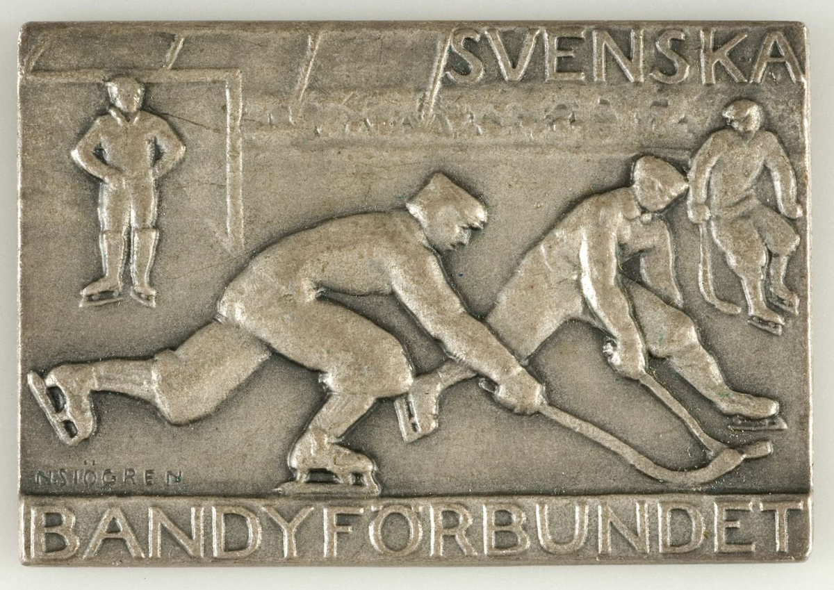 Rektangulär plakett av metall, silverfärgad. Vunnen i bandymatch. Motiv på framsidan: bandymatch samt texten: Svenska bandyförbundet. På baksidan texten: Div I Centrala 1965. Konstnärssignatur: N. Sjögren.
