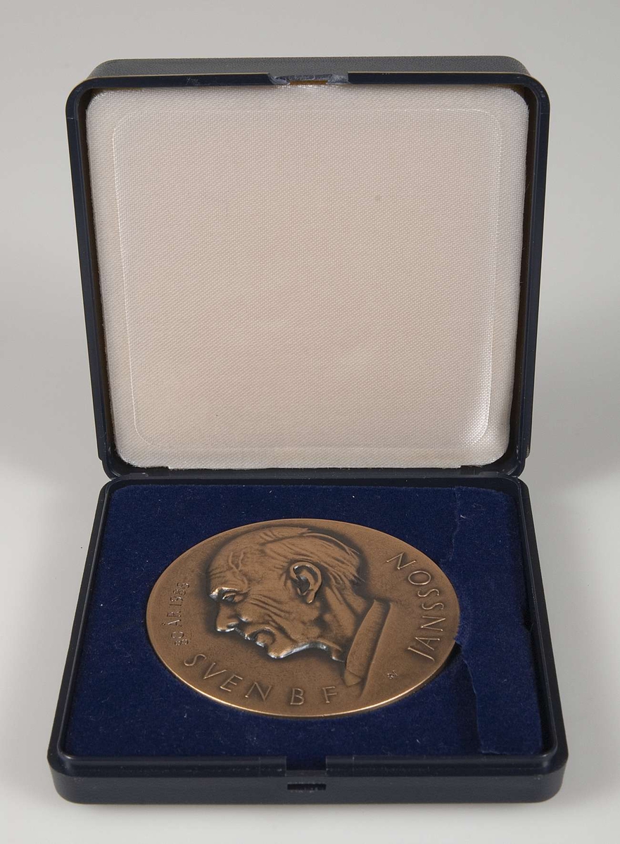 Rund plakett av brons. Tillhörande blå ask. Plakettens motiv på ena sidan: Manshuvud i profil med texten: Sven BF Jansson 80 år 1986. På baksidan: Motiv från runsten. Text: Runforskaren Riksantikvarien Run-Janne.