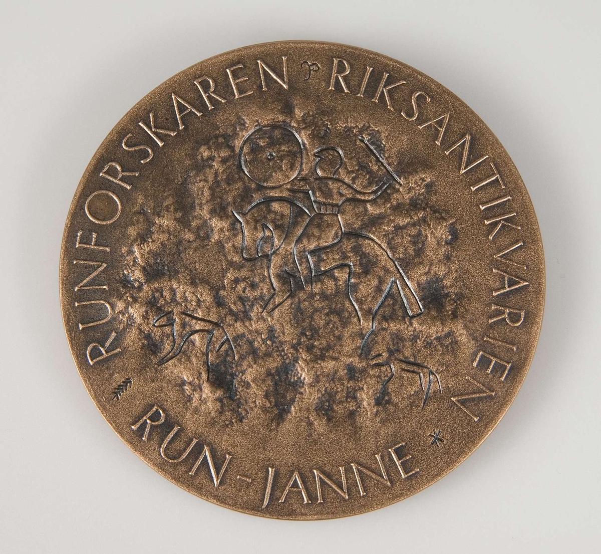 Rund plakett av brons. Tillhörande blå ask. Plakettens motiv på ena sidan: Manshuvud i profil med texten: Sven BF Jansson 80 år 1986. På baksidan: Motiv från runsten. Text: Runforskaren Riksantikvarien Run-Janne.