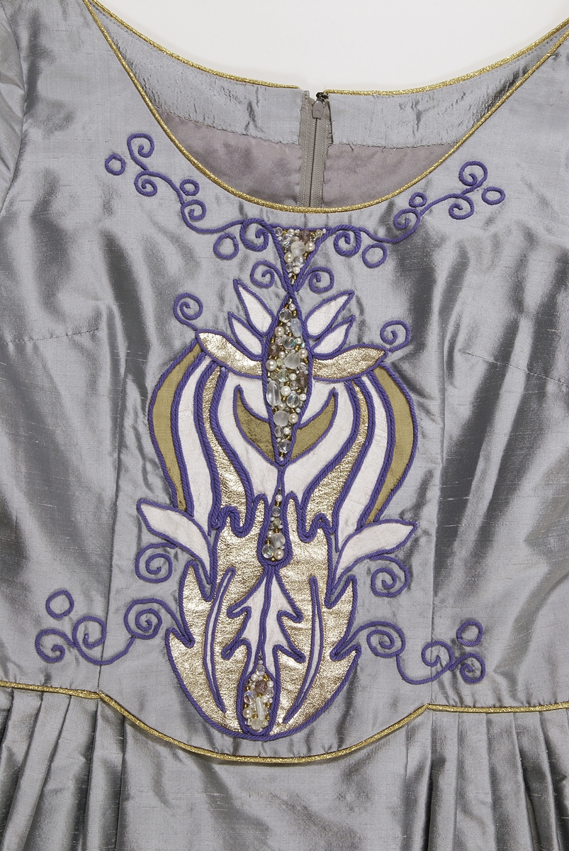 Klänning i grått siden. Figursytt liv. Broderier på framstycket med inslag av pärlor, lila garn och applikationer i guldfärgat och ljusgrått siden. Urringad hals kantad med guldband. På ryggsidan dragkjedja och hyska. Klänningen har smala långa ärmar som är klockade vid handlederna med infodringar av guldfärgat siden, kantat med guldband. Kjolen är utställd och har lagda veck i midjan. Längst ner på klänningen finns en cirka 30 centimeter bred våd av guldfärgat siden. Ovanför våden ett guldfärgat band. Klänningen är fodrad med grått fodertyg.