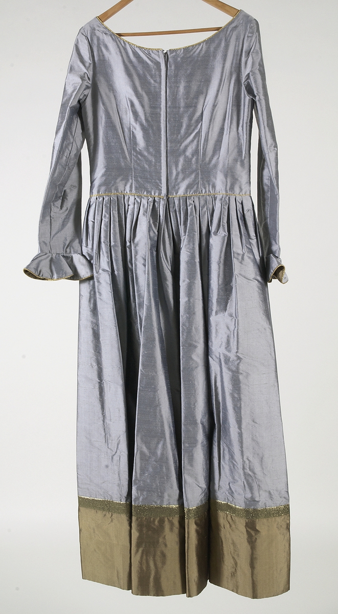 Klänning i grått siden. Figursytt liv. Broderier på framstycket med inslag av pärlor, lila garn och applikationer i guldfärgat och ljusgrått siden. Urringad hals kantad med guldband. På ryggsidan dragkjedja och hyska. Klänningen har smala långa ärmar som är klockade vid handlederna med infodringar av guldfärgat siden, kantat med guldband. Kjolen är utställd och har lagda veck i midjan. Längst ner på klänningen finns en cirka 30 centimeter bred våd av guldfärgat siden. Ovanför våden ett guldfärgat band. Klänningen är fodrad med grått fodertyg.