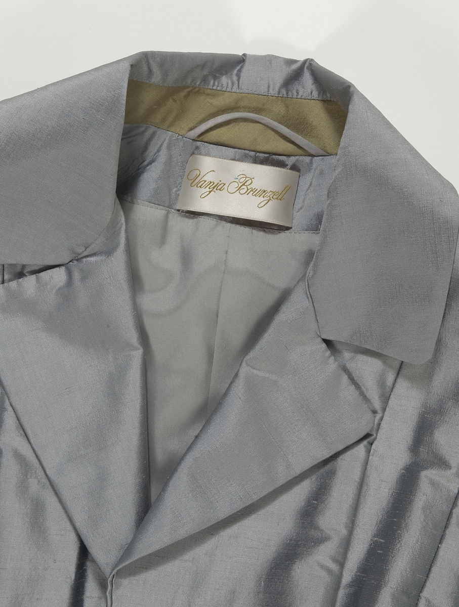 Kappa i grå siden. Kappan, som är formsydd i överstycket, har en utställd kjoldel  med lagda veck runt midjan. Vid halsen en krage med slag. Kappan har långa ärmar med en slits vid handleden. Vid slitsen kant av guldfärgat siden.