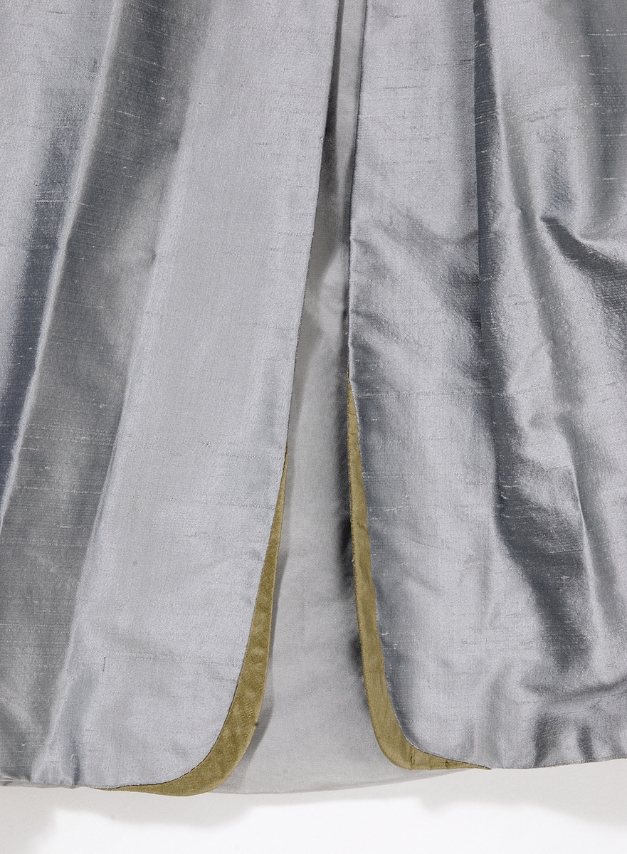 Kappa i grå siden. Kappan, som är formsydd i överstycket, har en utställd kjoldel  med lagda veck runt midjan. Vid halsen en krage med slag. Kappan har långa ärmar med en slits vid handleden. Vid slitsen kant av guldfärgat siden.