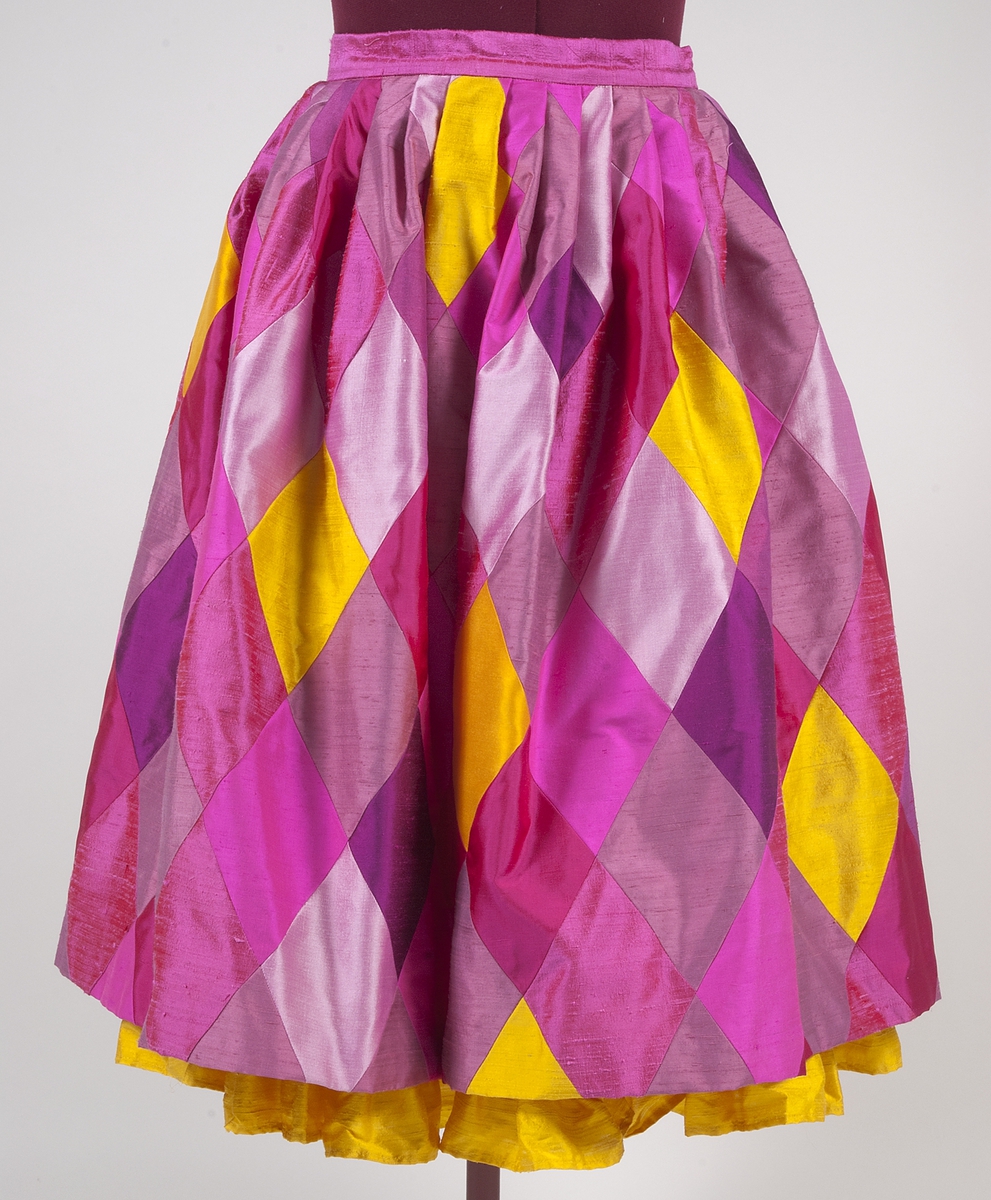 Kjol med rombformade sammansatta bitar i rött, flera nyanser av rosa och gult siden. Kjolen har lagda veck i midjan så att den får vidd. Linning av rosa siden. Kjolen stängs med en rosa knapp och blixtlås på där bak. Kjolen är fodrad med ett rosa fodertyg. Längst ner på fodret finns en 15 cm rynkad våd av gult siden som syns nedanför kjolens kant.