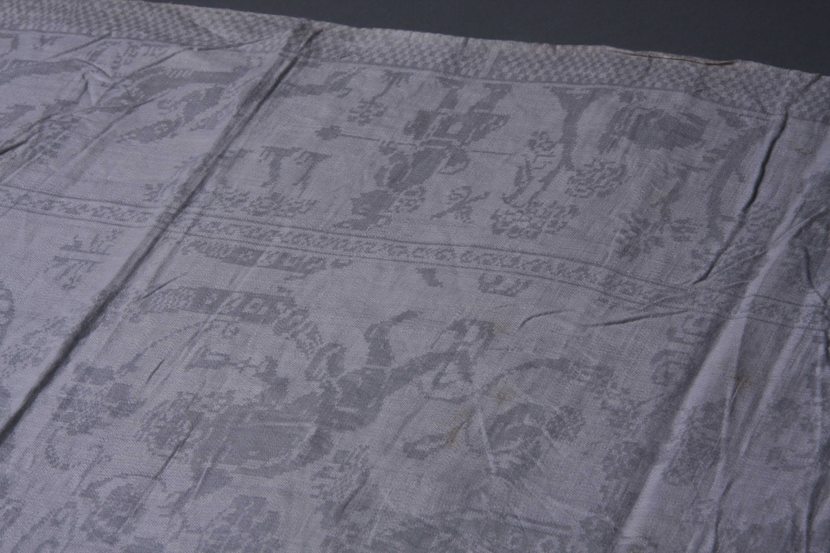 Hvit lindamask med sjakkbrettbord  på begge langsider. Innenfor en bord med jegere til fots, hunder og vilt.På kortsidene ryttere, og i to av hjørnene et slott.
Midtpartiet opptaes av båndlignenede ranker som går ut fra et firkantet felt midt på servietten, båret av to flyvende puttier. Mellom rankene Pan og puttier.
Smal fall på kortsidene.