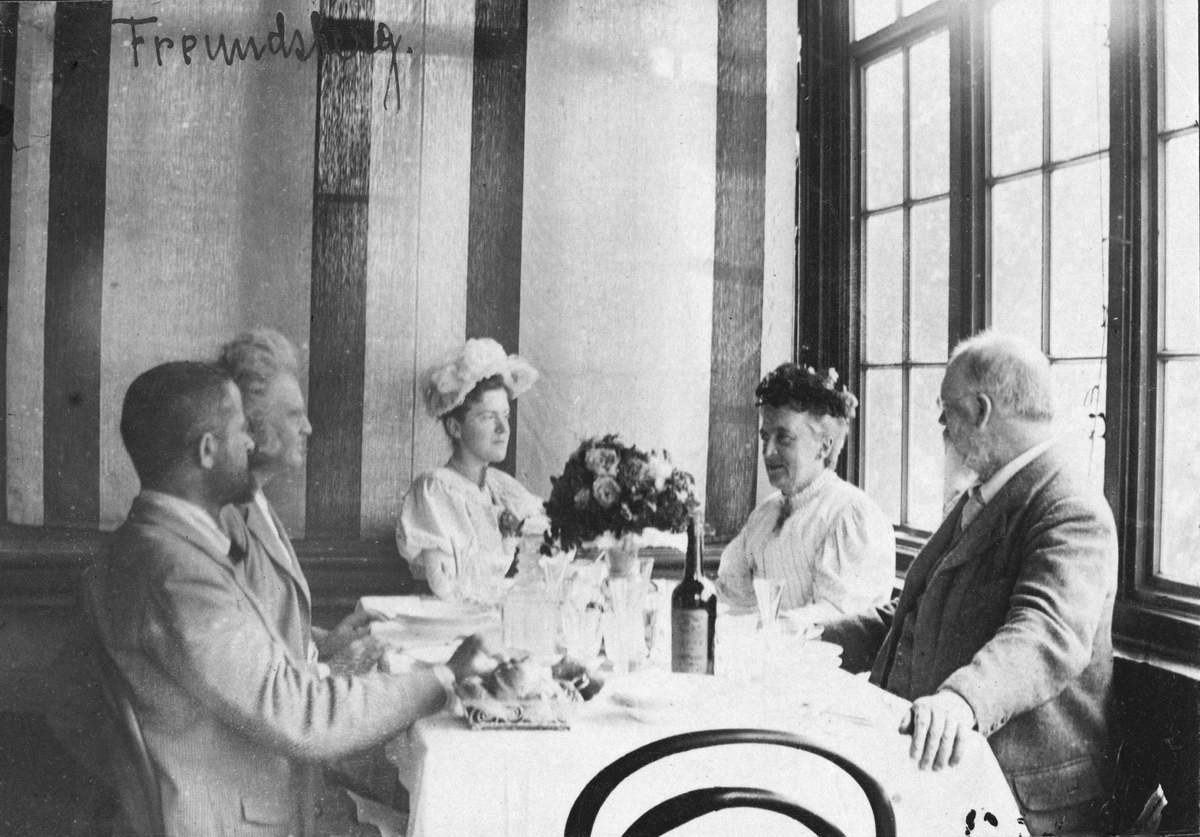 Fotografi fra spisesal; ved bordet sitter to damer og tre herrer; de ser på hverandre.