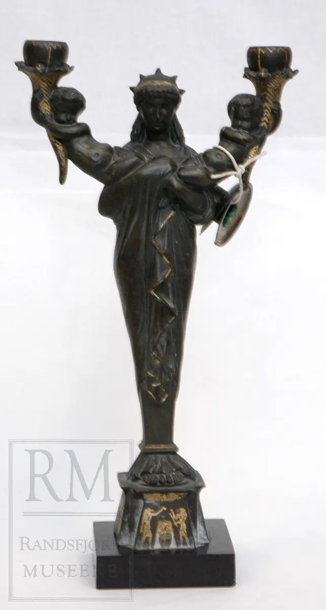 Kvinnefigur på sokkel, med ett barn i hver arm. Barna holder hvert sitt horn (?) med lyseholder. Noe forgylling. Løse lysmansjetter. b) mangler sokkel.