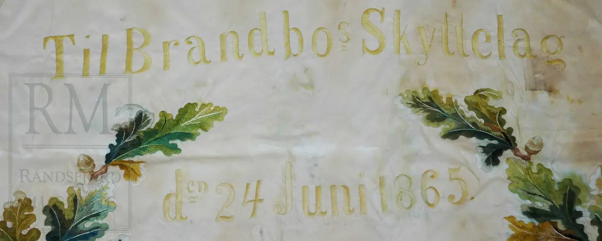 Fane av hvit silke. Løpegang på den ene siden har revnet. Brodert tekst, samt eikebladskrans med sløyfe omkring to gevær med påhengende krutthorn.