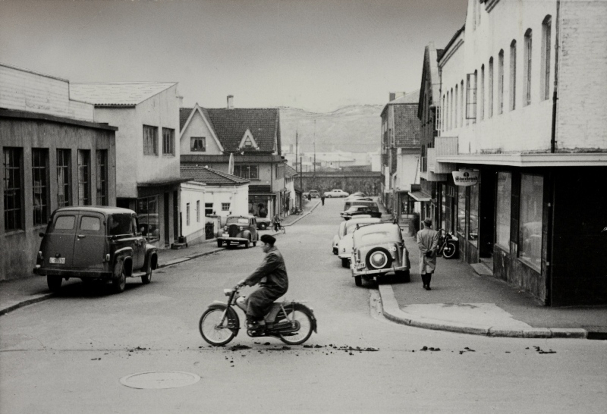 Sandnes sentrum i 1958. Fotografen står i Storgaten og ser nedover Erling Skjalgsonsgate mot St. Olavsgate og høybanen. Konfeksjonfabrikken ligger bak til høyre i bildet. Tidligere var den i det fremre bygget til høyre. Bildet er tatt for sammenligning med et fotografi tatt av samme sted rundt år 1900.