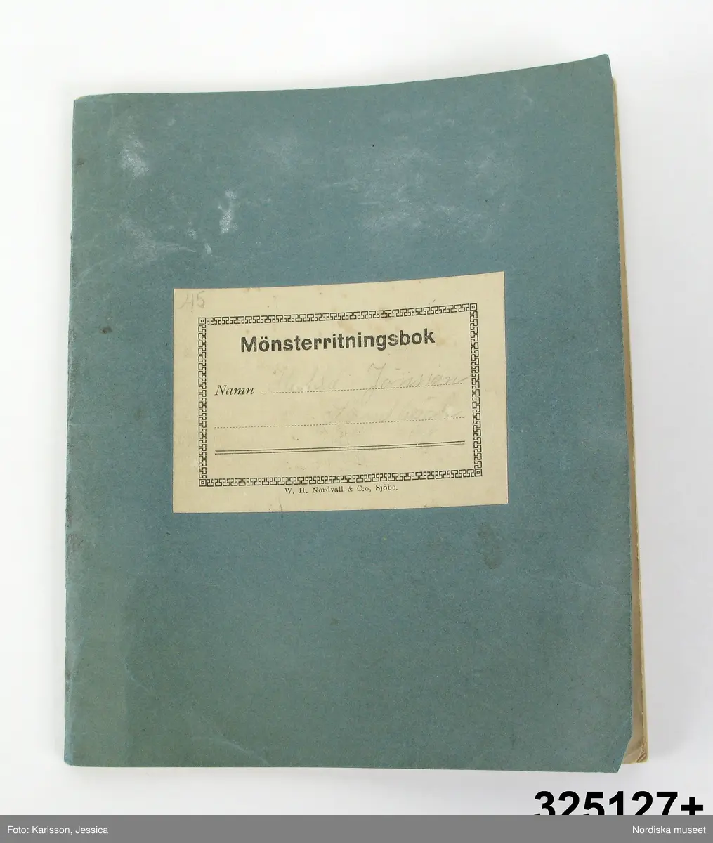 Häfte med blått omslag med etikett på framsidan med tryckt: Mönmsterritningsbok samt påslkrift svagt med blyerts:" Hulda Jönsson, Sandbäck." Tryckt med liten stil: W.H.Nordwall & Co, sjöbo.
Innehåller 26 st 2-mm rutade blad av vilka 15 har anteckningar, solvnotor mm. till vävnader i blyerts och bläck samt färgkrita, bl.a "Gardin nota, Trasmatta, Gardin i myggtjäll, Bomullstyg, Handukar i gåsögon, Handdukar i korndräll, Daldräll, Rosengång, Matta i bomull, Tramatta där varpen bildar fasonerna. Trasmatta med bård på längden." Några verkar avskrivna ur tryckt bok. Dessutom udda mönsterfigurer bl.a opphämtamönstret till täcke/duk inv.nr 325137.
Anm. Fläck på baksidan.
Två tygprover, ett löst 7 x 6,5 cm, och ett fäst med knappnål 11 x 5 cm, båda vävda i gles tuskaft med tunt tvinnat vitt bomullsgarn. Dessutom 13 lösa papperslappar i olika storlek och sort varav några hopfästa med knappnålar. Med anteckningar, solvnotor mm. till vävnader och även ett knyppelmönster till en uddspets.
Hulda Jönsson var född 1885 i S. Åsums sn, dotter till statdrängen Jöns persson, arbetade som piga på en gård i Sandbäck i S.Åsum, där hon lärde sig väva. På 1920-talet vävde hon på beställning åt privatpersoner och Hemkultur i Malmö, från 1930 trasmattor som såldes genom "Kooperativa i Sjöbo". Se textilier och vävböcker inv.nr 325 125 - 137.
Se vidare bilaga och D.408
/Ingrid Frankow 1999/
/Berit Eldvik 2011-03-02