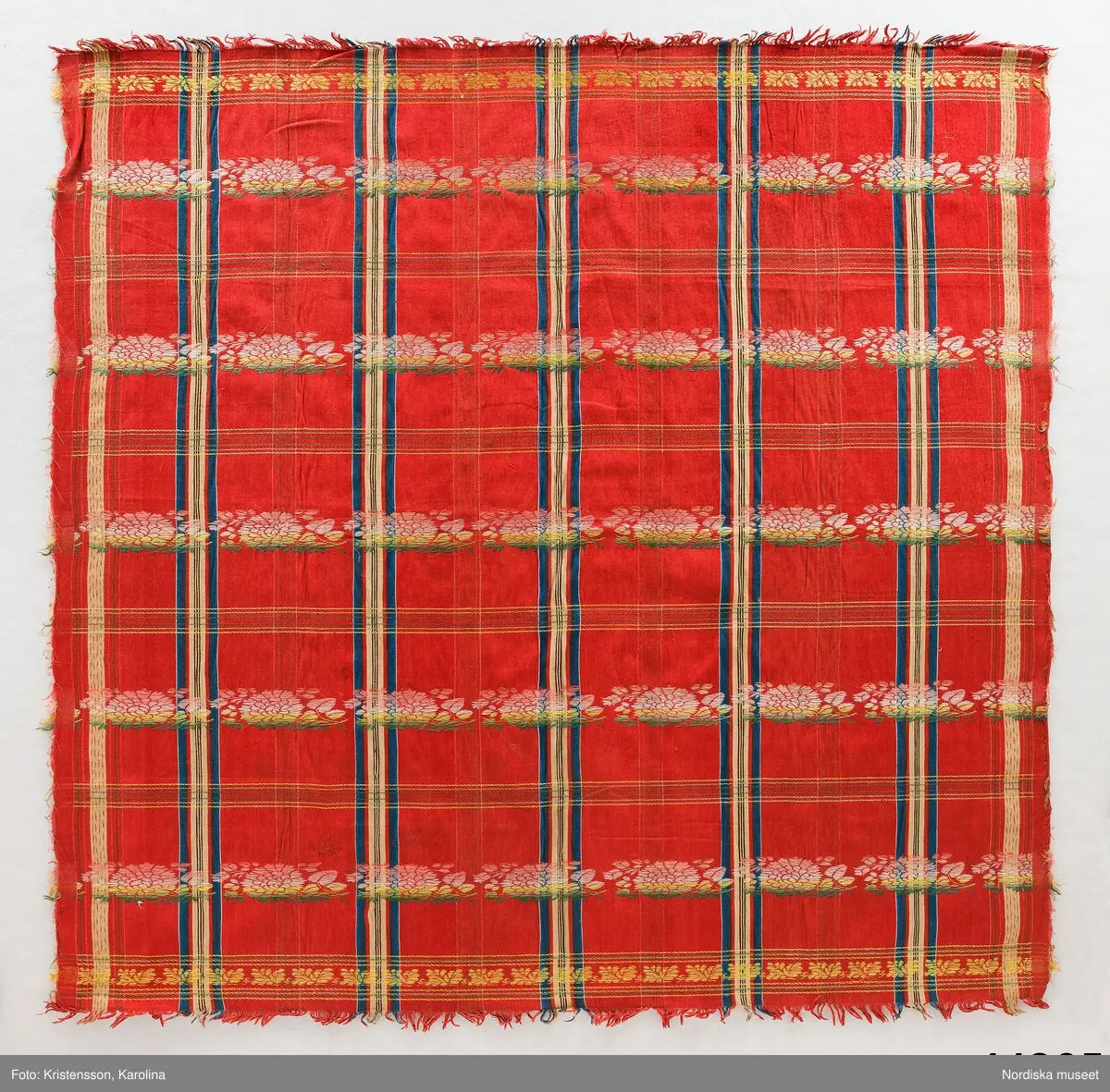 Kvadratiskt halskläde av  halvsiden.  Storrutig  på klarröd botten i satin  med  ränder i schatterande blått, beige, rött och brunt och med jacquardvävda tvärgående blombårder  i rosa, grönt  och gult silke med smala gula  kantbårder. Runtom upprispad kort frans. I ena hörnet en rund svart stämpel med svårläst text: Stockh. Det är Stockholms hallstämpel jfr. likadan t halskläde 44.265 som har en tydligare stämpel  från 1830-talet.
I mycket gott skick.
Troligen en brudschal. Se samma mönster i mörkblått 91.624 från Unnaryd i Småland.
/Berit Eldvik 2012-01-27