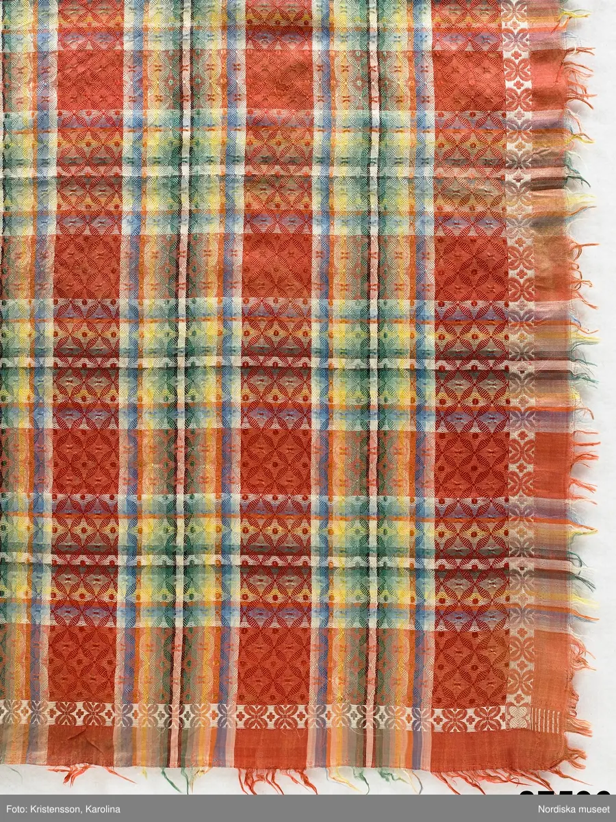 Kvadratiskt halskläde av siden, orangeröd botten med rutning i gult, grönt, ljusblått och vitt samt en smal ytterbård med stiliserad blomma längs 2 sidor i vitt och längs de andra i gult. Botten mönstervävd i ett bindemönstermönster med kryss och små stjärnor, i ytterkant utanför bården slät tuskaft. Uppsrispad kort frans runt alla sidor. I ena hörnet en rund svart hallstämpel med texten. Stockholm 1825 och i mitten St. Eriksbild. Troligen vävd i en skaftvävstol.
Typen var vanlig på 1820-talet och finns från flera håll i landet bl.a 98530 från Äppelbo och 93669 från Malung  i Dalarna. även 44279 från Västmanland och 91632, 152104, 162623 från Småland.
I fint skick
/Berit Eldvik 2011-11-23