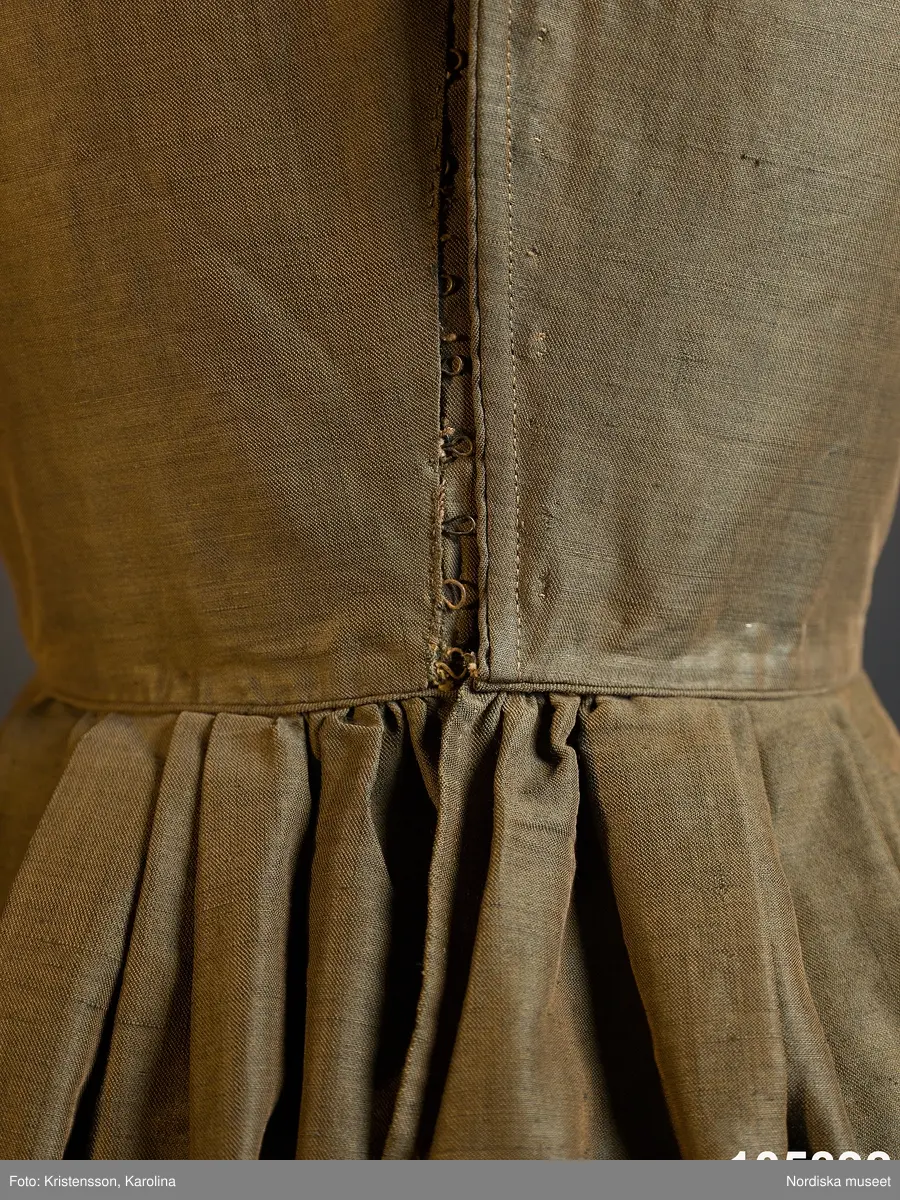 Hel klänning i 1830-talets modesnitt, av fint halvylletyg i kypert med varp av ljusbrunt bomullsgarn och inslag av grönt tunt glansigt redgarn som ger en grönskimrande yta. Livet går ned nästan till midjehöjd, figursytt med 2 framstycken med bakåtflyttade sidsömmar, ett ryggstycke med 6 inprovningar fördelade i två grupper. Vid båtringad halsringning, knäppning med tätt sittande hakar och hyskor. Isydda långa ärmar svängda med 2 sömmar  med 3 veck vid armbågen. Ärmsprund knäppt med hyska och hake samt 4 cm uppslag med rundade hörn. Upptill på ärmen besättning av tyget med stora rundade uddar kantade med klargrön bomullsfrans.  Passepoaler av tyget i alla sömmar. Livet fodrat med skotskrutig bomullslärft i rött, grönt, gult och vitt och i ärmarna ett smalrandigt bomullstyg i blått och rosa. Kjolen sydd av  5 våder, lagda veck runtom i midjan. sprund mitt fram. Uppsytt veck  en decimeter från fållen. Skoning med brun bomullslärft. Handsydd.
Har burits 1838 som ungmorsklänning, alltså av bruden dagen efter bröllopet, av Maria Margareta Mårtensdotter från Kysings, Vall sn på Gotland. Möjligen tillsammans med sidenhalsklädet 105385.
Anm. I  gott skick.
OBS Klänningen sitter bakochfram på fotona, knäppningen är fram.
/Berit Eldvik 2011-06-13