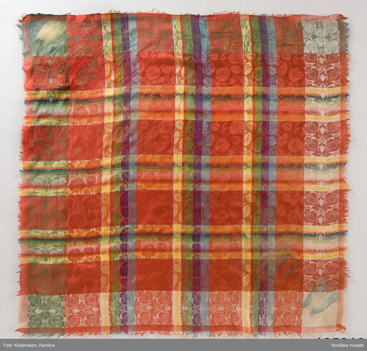 Kvadratiskt halskläde av siden, klarröd botten med rutor i grönt, ljusblått, ljuslila och gult med ett yttäckande invävt mönster, troligen med jacquard, med slingrande rosor. Kantbård med "päronlika" blommor/frukter som på 2 mötande sidor är gröna och på de 2 andra vita. Upprispad kort frans på alla sidor.
I ena hörnet en tydlig svart rund stämpel med texten: Stockh. hallstemp 1839.
Anm. Stor fläck i två av hörnen.
/Berit Eldvik 2011-11-11