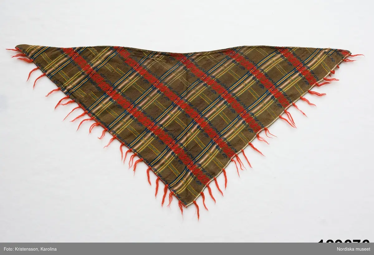 Trekantigt halskläde av troligen jacquardmönstrad bomullssatin.  Grönbrun botten randig i  ljusare grönt, gult och blått med invävd bladslinga i bottenfärgen. Tvärgående mönsterbårder i rött och blått  samt smalare i gult och grönt, vilket ger ett rutigt  tyg. Tvärbårderna i inslagsflotteringar.   Tyget har 2 materialskarvar. I ytterkanterna iknuten gles frans av rött ullgarn.Fodrad till större delen med ett grön/brunrutigt bomullstyg, snibben bak ofodrad.
 Tillverkad av delar från ett kvadratiskt halskläde.
/Berit Eldvik 2012-02-09