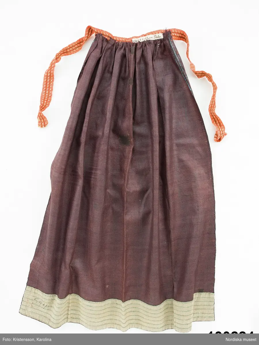 Förkläde av mörklila tunn yllekypert av kamgarn med starkt glättad yta, s.k. rask eller chalon. har fått ett sekundärt stämpeltryckt mönster med oljefärg, smala dubbla  ränder  i klarblått och mellan ränderna rader av  "rosor" dvs. mönsterfigurer i vitt och rött. Rynkat i midjan med ett påsytt  3 cm brett midjeband i ylle smårutigt  i rött, grönt och vitt av den sort som kallas Malsband dvs. Malungsband. Nederkanten inskodd med  smalrandigt halvylletyg i grått och grönt/ljusgult. Handsömnad.
Rask eller chalon var ett köpetyg som användes till högtidsförkläden. Här har man låtit en lokal tryckare mönstra tyget, vilket är ovanligt på denna typ av förkläde. 
erit Eldvik 2012-03-29