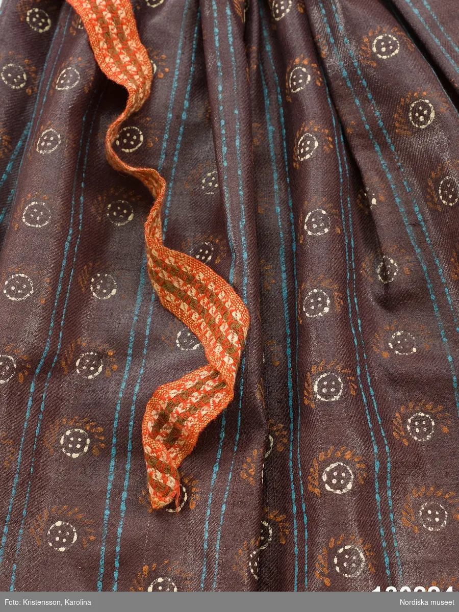 Förkläde av mörklila tunn yllekypert av kamgarn med starkt glättad yta, s.k. rask eller chalon. har fått ett sekundärt stämpeltryckt mönster med oljefärg, smala dubbla  ränder  i klarblått och mellan ränderna rader av  "rosor" dvs. mönsterfigurer i vitt och rött. Rynkat i midjan med ett påsytt  3 cm brett midjeband i ylle smårutigt  i rött, grönt och vitt av den sort som kallas Malsband dvs. Malungsband. Nederkanten inskodd med  smalrandigt halvylletyg i grått och grönt/ljusgult. Handsömnad.
Rask eller chalon var ett köpetyg som användes till högtidsförkläden. Här har man låtit en lokal tryckare mönstra tyget, vilket är ovanligt på denna typ av förkläde. 
erit Eldvik 2012-03-29