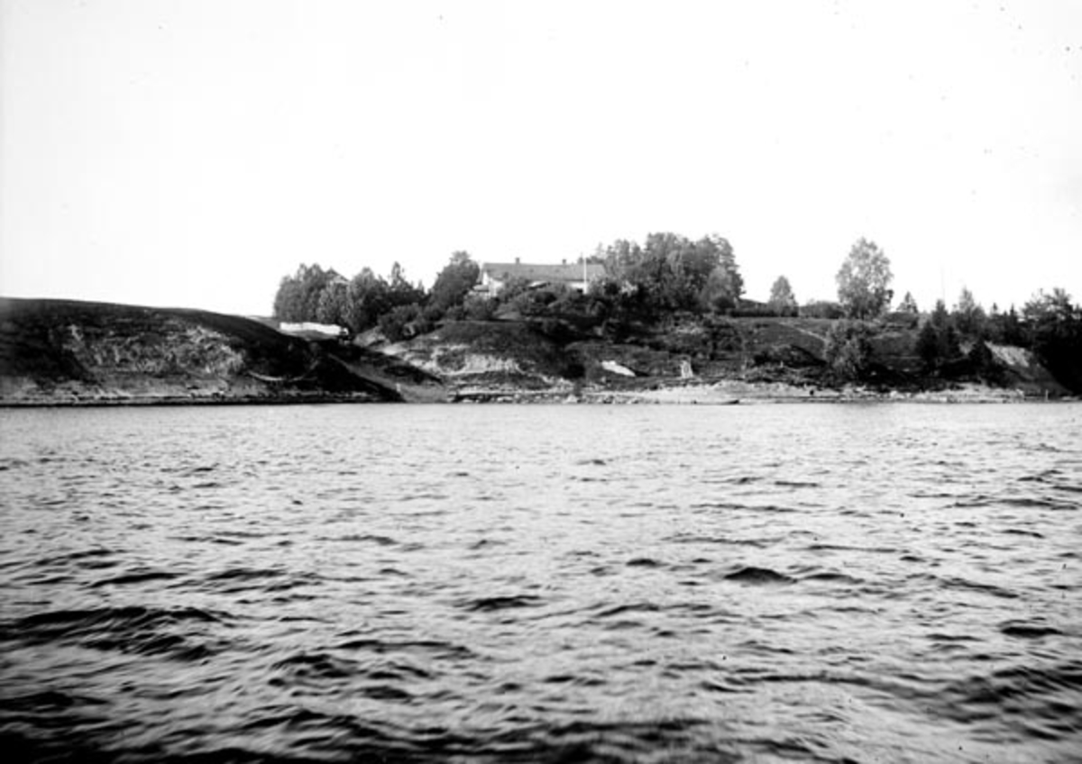 Garden Solberg i Sørum kommune på Romerike.  Fotografiet er tatt fra den andre sida av Glomma, over vannet, mot bergryggen ved elva, der taket på hovedbygningen og flaggstanga rager over vegetasjonen i hagen.  På denne eiendommen bodde Adolf Brede Øvergaard (1854-1917), som var tømmermåler.