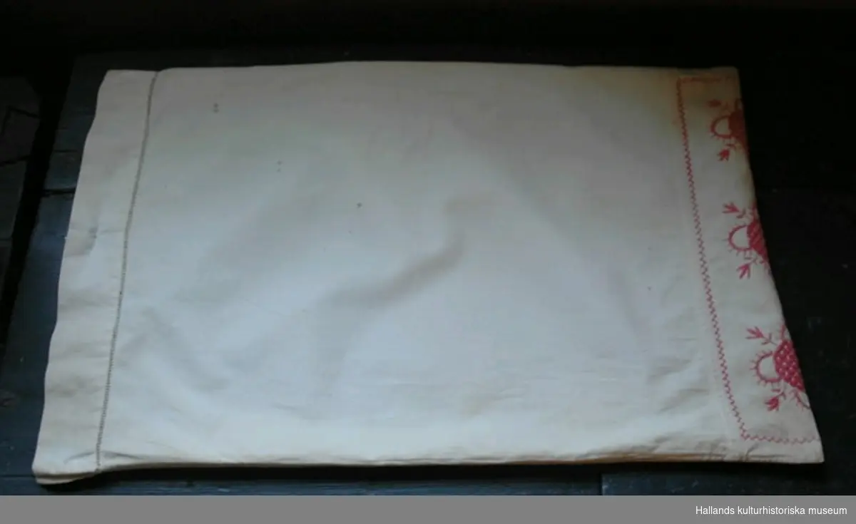 Örngott av vävt bomullstyg med klädda knappar i öppningen. Kanten utmed öppningen ör sydd med hålsöm. Ena sidan broderad med röd hallandssöm  Broderade initialer: "CB" i jugendstil.