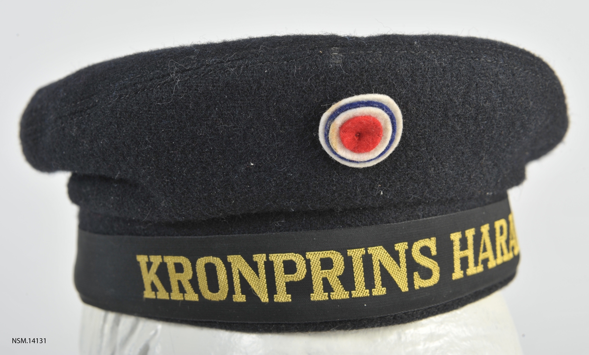 Rund matroslue i vadmel med navnet Kronprins Harald rundt bremmen.