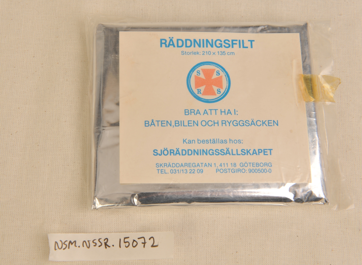 Redningsduk av tynn aluminium foldet sammen og lagt i en klar plastikkpose. Opplysninger på kartong i posen.