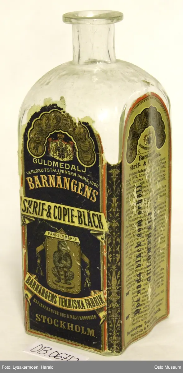 Flasken har etikett over tre sider. Den har mørk beige bunnfarge, og trykk i sort, blått, rødt og gull. Tegning av en bjørn som rører i en bøtte med blekk under teksten "FABRIKSMÄRKE".