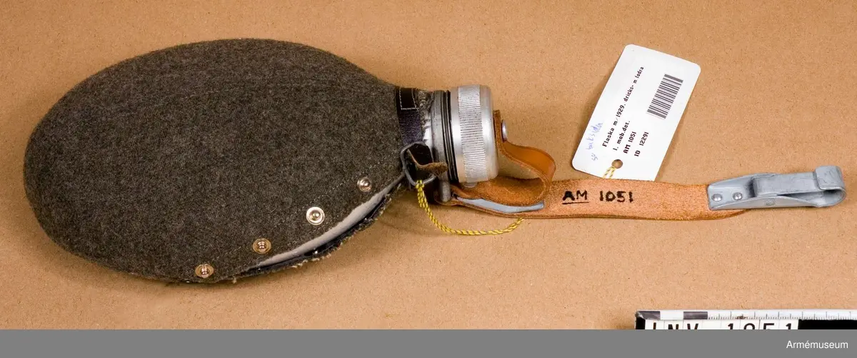 Samhörande nr är 1017-1059, 1062-1063, 1072-1073.Dricksflaska m/1929, m fodral.Med skruvlock, rem och fodral av ylle. Njurformad med något konkav insida (den sida som bärs mot kroppen) och konvex sida utåt. Vid flaskans hals finns en fastgörningsanordning för dels en rem för att hålla fast locket med, dels en längre rem med karbinhake för fastsättning i ryggsäcken. Locket skruvas fast vid flaskans hals. För "greppet" är lockets kant refflad. Skydd av gråbrungrönt yllefodral. Fodralet är knäppt med fyra tryckknappar. Runt "halsen" är fodralet skott på rätsidan med ett brunt läderband som knäpps med ett litet metallspänne med  ett torn. Se AM 1052. Flaskan tillhör mobiliseringsutrustning vid museet.