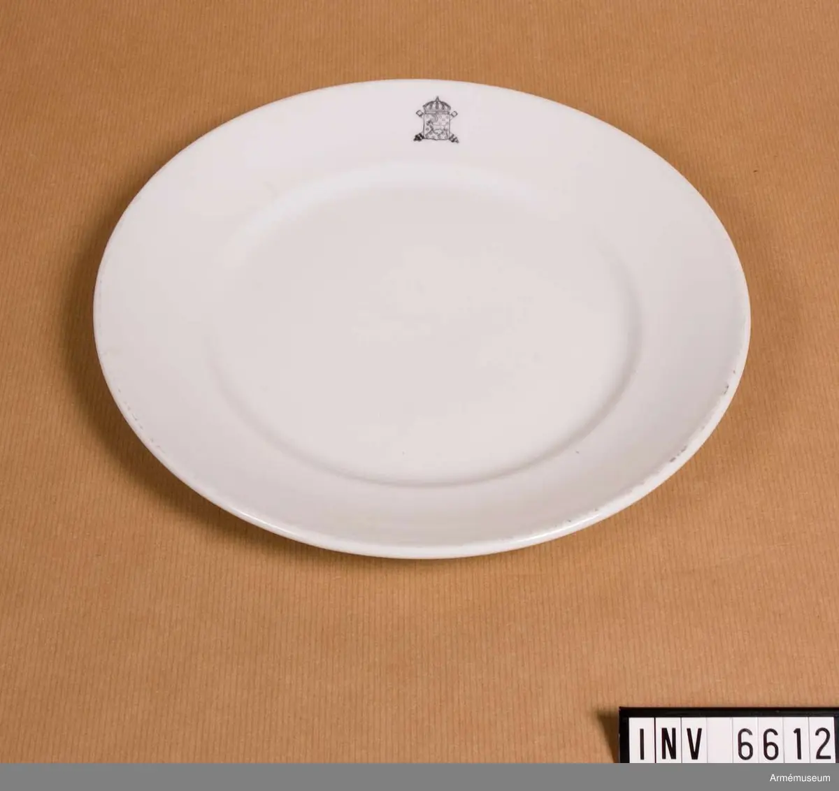 Flat tallrik, Norrbottens artillerikår A 5, 1925-1951. I vitt  porslin med A 5:s vapen i svart. På undersidan är fatet märkt:  RÖRSTRAND Sverige 492, kronstämpel som firman började använda  år 1884.