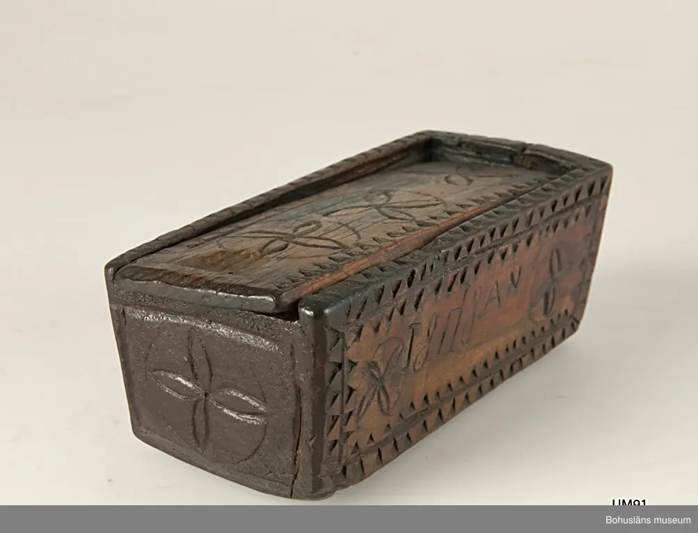 Rektangulär låda med skjutlock och något snedställda kortsidor.
Skuren dekor bestående av rosetter, kors samt uddsnitt längs kanterna.
På en av långsidorna är årtalet "1848" och initialerna "AN" inskurna.
Inuti ligger en äldre etikett från ca sekelskiftet.
Kan vara en friargåva.

Litteratur:
Gjaerder, Per, Esker og tiner, C. Huitfeldts förlag, Oslo, 1981, s. 41.
Knutsson, Johan, Friargåvor, Nordiska museets förlag, Stockholm, 1996.

Ur handskrivna katalogen 1957-1958:
"Trälåda
1870-1887
Bottenmått: 14 x 5,5 H. 5. Trä med skuren ornering. Föremålet helt. Bohusl."

Lappkatalog: 84