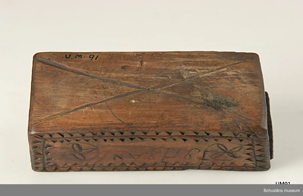 Rektangulär låda med skjutlock och något snedställda kortsidor.
Skuren dekor bestående av rosetter, kors samt uddsnitt längs kanterna.
På en av långsidorna är årtalet "1848" och initialerna "AN" inskurna.
Inuti ligger en äldre etikett från ca sekelskiftet.
Kan vara en friargåva.

Litteratur:
Gjaerder, Per, Esker og tiner, C. Huitfeldts förlag, Oslo, 1981, s. 41.
Knutsson, Johan, Friargåvor, Nordiska museets förlag, Stockholm, 1996.

Ur handskrivna katalogen 1957-1958:
"Trälåda
1870-1887
Bottenmått: 14 x 5,5 H. 5. Trä med skuren ornering. Föremålet helt. Bohusl."

Lappkatalog: 84