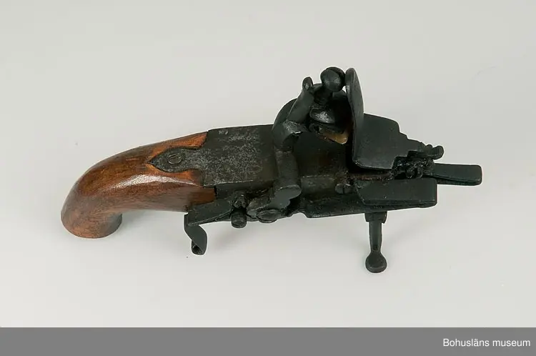 Ur handskrivna katalogen 1957-1958:
Elddon, "1800".
L. 22, H. c:a 13 cm; ett flintlås monterat m. handtag av trä och 2 fötter av järn; på sidan en lucka på gångjärn, m. regel, på denna (d.v.s, på luckan) en ljushållare av järn. Helt.

Lappkatalog: 90