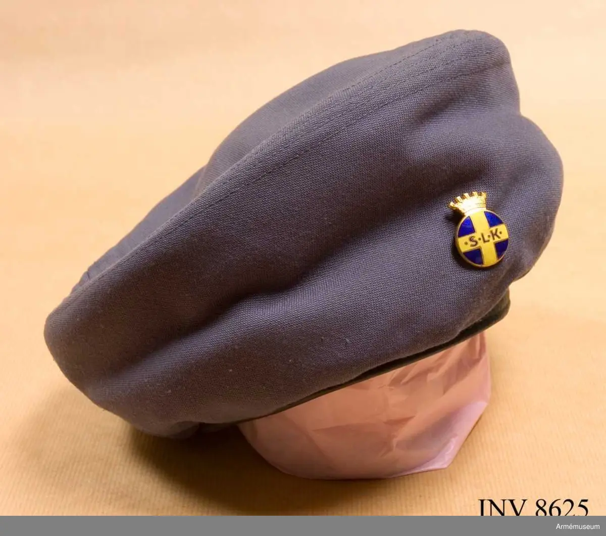 Samhörande gåva 8617-8626.
Tillverkad av samma tyg som föreningsdräkten. Den bärs enligt regler för civil kvinnlig huvudbonad. Skall alltid medtagas på övningar, skolor och liknande. Baretten har och skall ha lottabroschen över vänster öga ca. Källa SLK Värt att veta... 1970 sid 11-12.